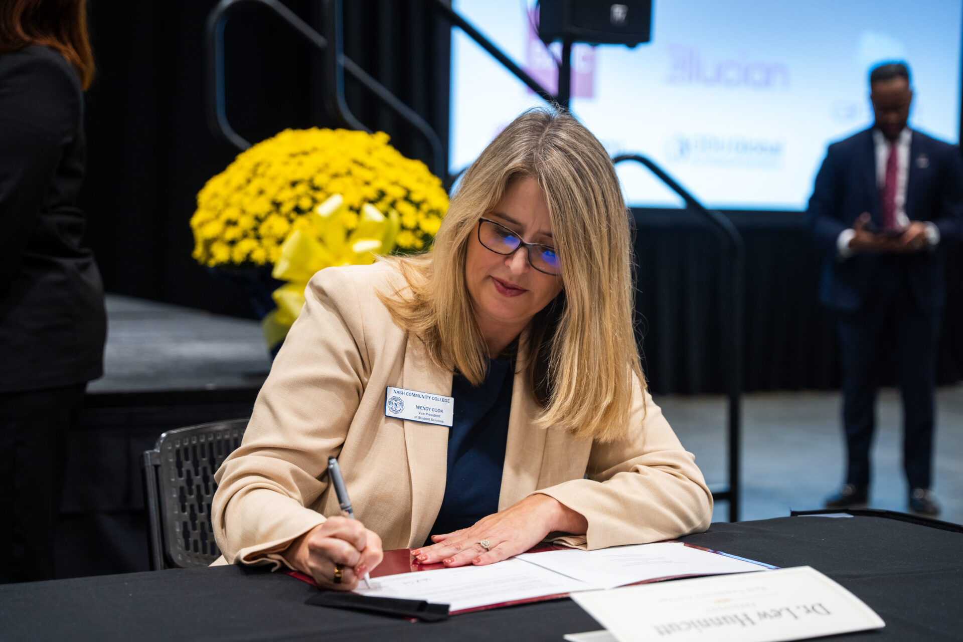 A person with glasses and a name badge signs a document at a table during an event, with a large yellow floral arrangement and a screen displaying logos in the background.