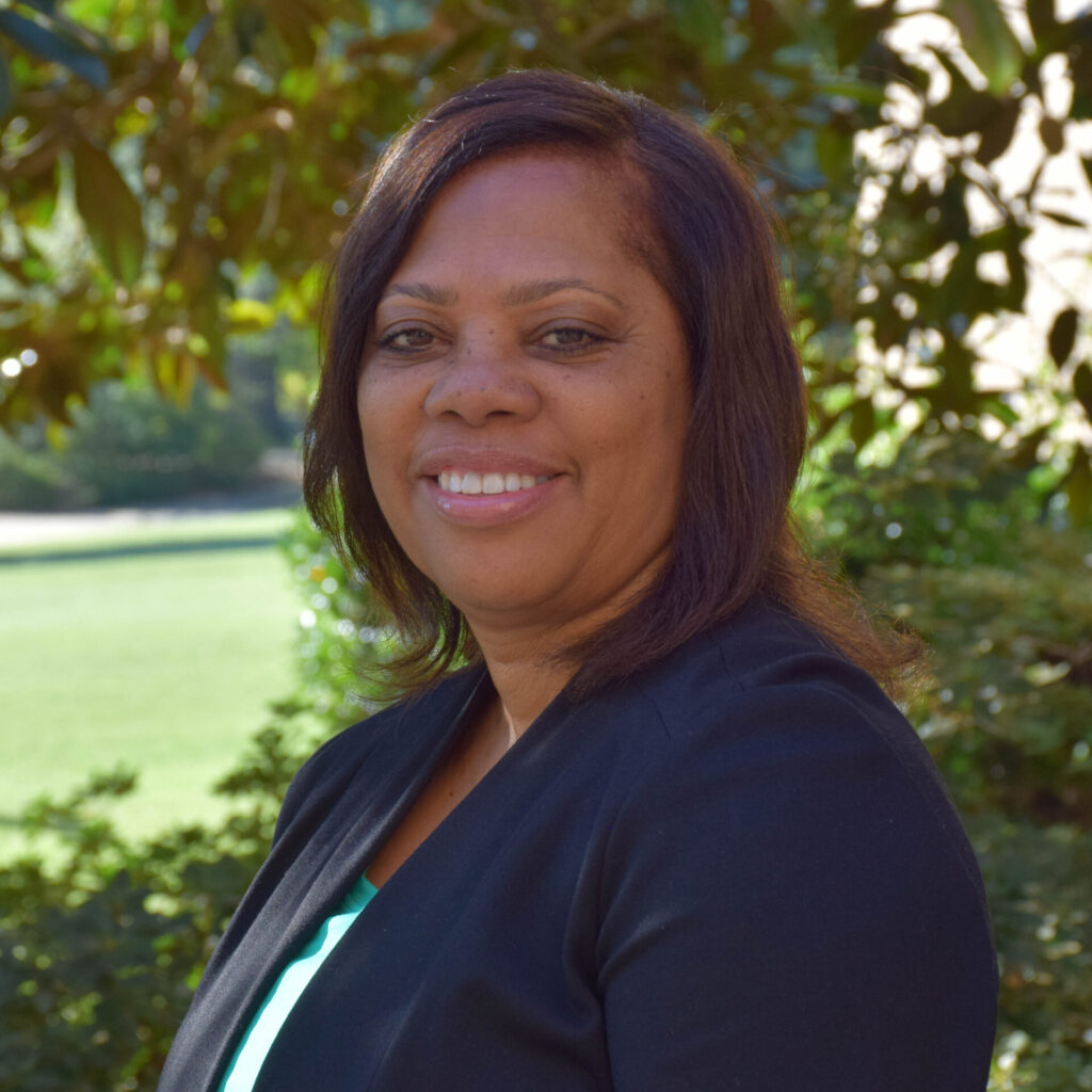 Brown skinned woman standing for a headshot with trees in the background.