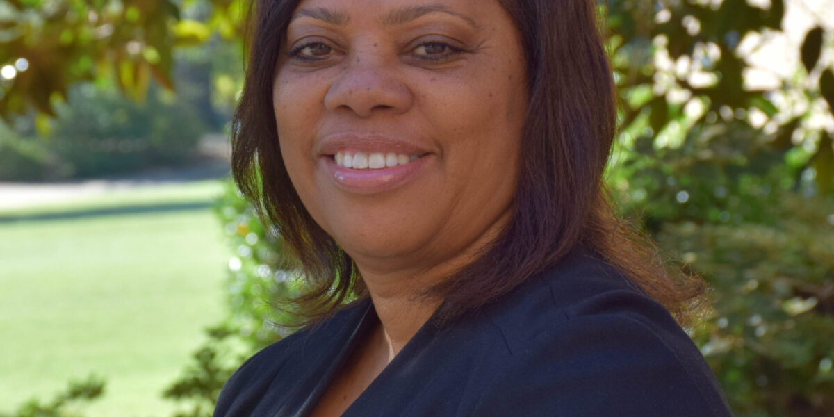 Brown skinned woman standing for a headshot with trees in the background.